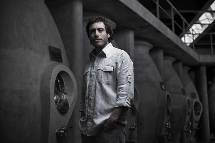 A man in a denim shirt standing in front of large cement tanks in a winery setting