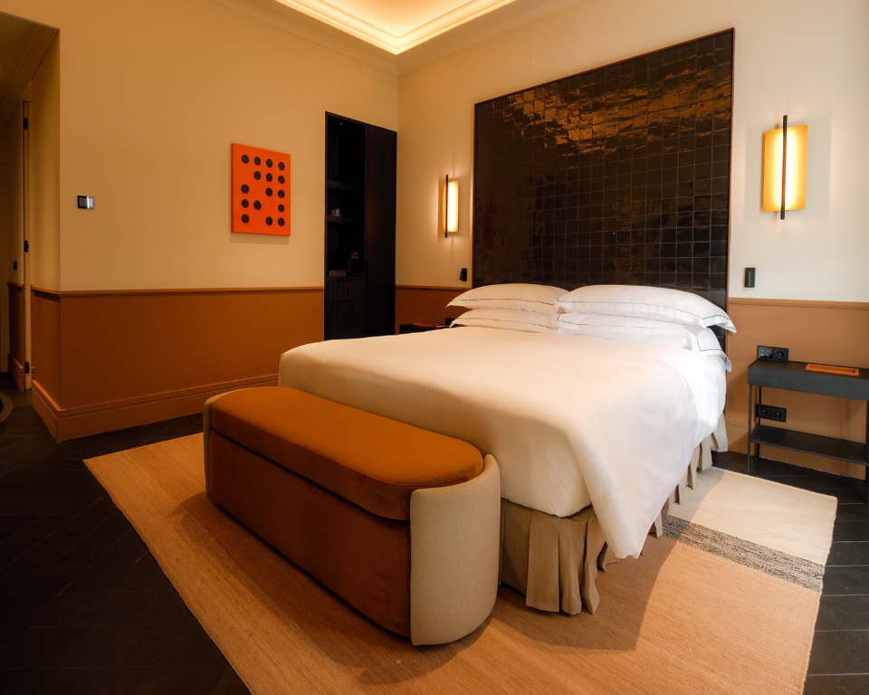A luxury hotel bedroom with white bedding, dark tiled headboard, warm wood paneling, modern wall sconces, and contemporary art