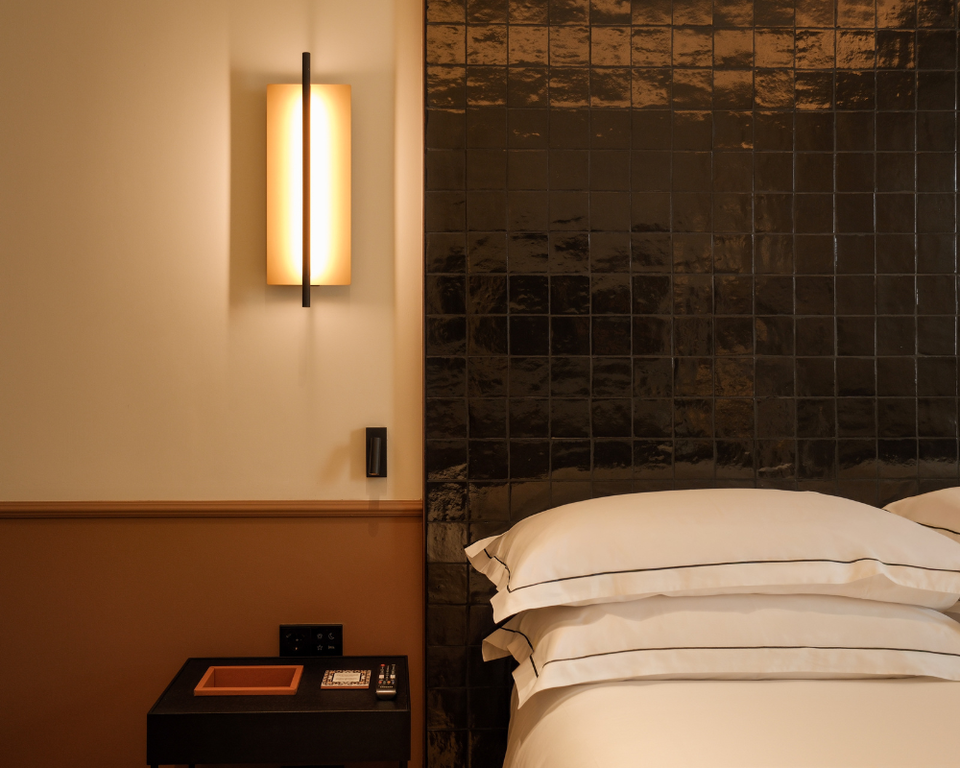 A hotel bedroom interior featuring a bed with white linens against a dark glazed tile wall, with a modern wall sconce and wooden nightstand; the sourc