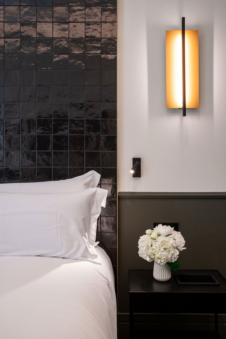 A luxury hotel guest room interior featuring a bed with white linens against a dark glazed ceramic tile headboard wall, with a modern wall sconce and