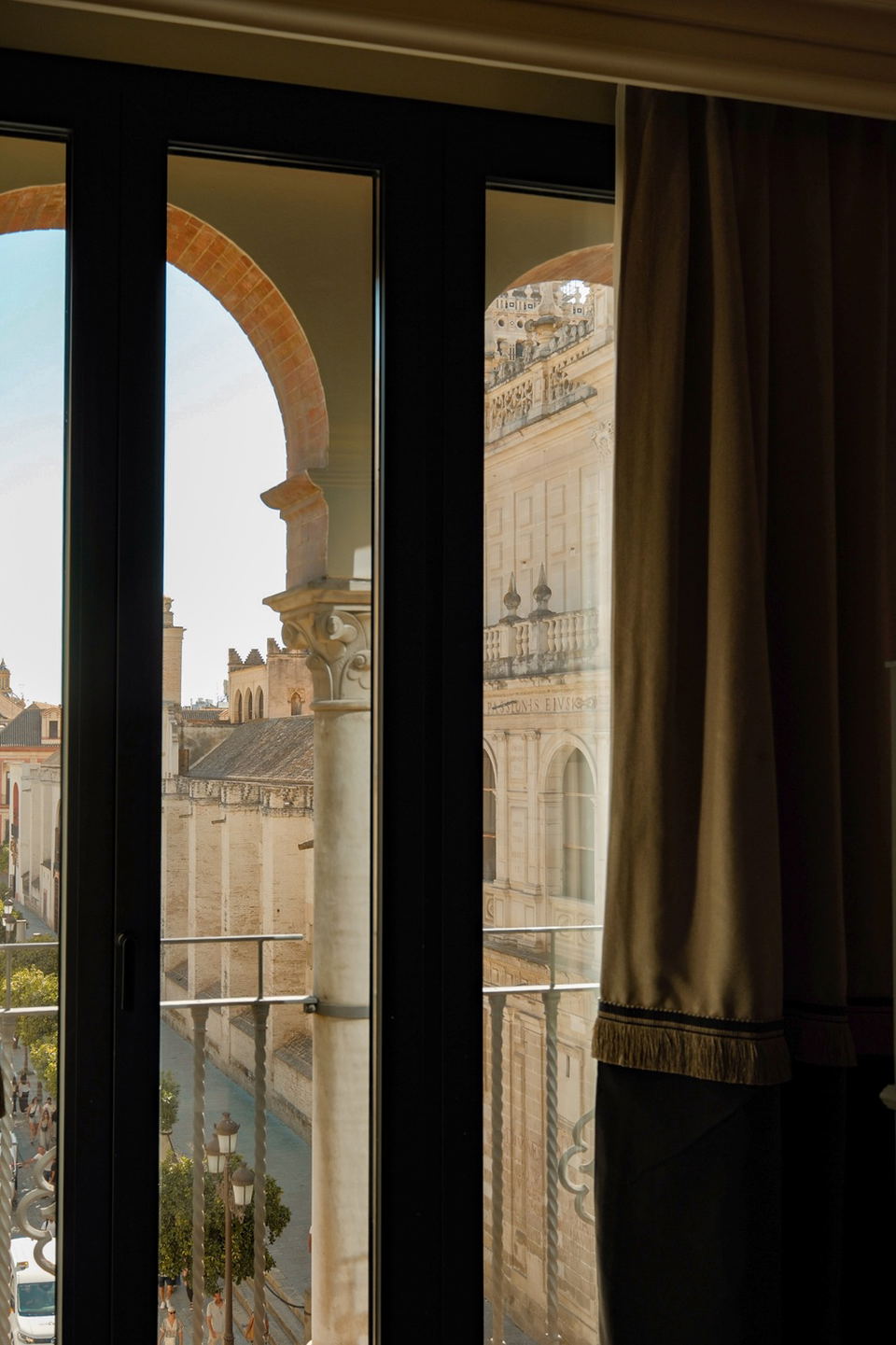 Interior view through glass doors/windows showing Seville Cathedral and La Giralda tower in the background, with elegant curtains framing the view; so