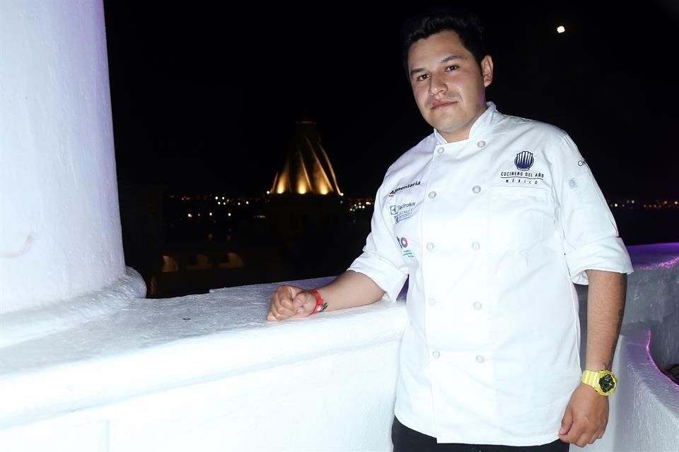 A chef in white chef's coat standing at a white wall/balcony at night with illuminated structures visible in the background