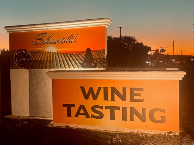 A Shanti Winery sign at sunset, featuring a vineyard scene with a meditating figure and the words "WINE TASTING."
