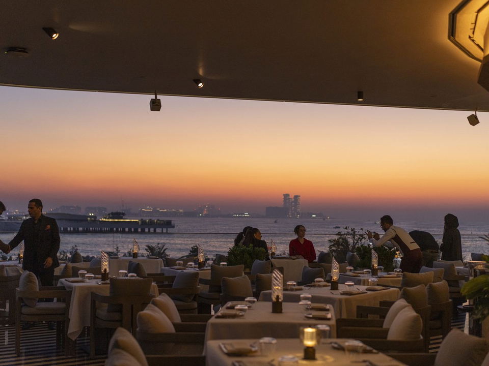 An upscale restaurant with a view of the sea at sunset, featuring elegantly set tables and staff attending to guests.