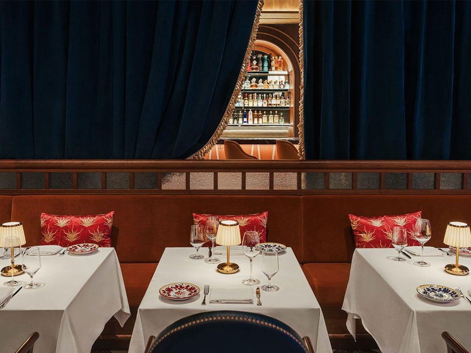 An elegant restaurant dining room with rust-colored banquettes, red patterned cushions, white-clothed tables, and a glimpse of a bar through blue