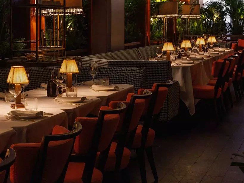 An elegantly set dining room with warm amber lighting, featuring small table lamps, white-clothed tables, rust-orange upholstered chairs, and
