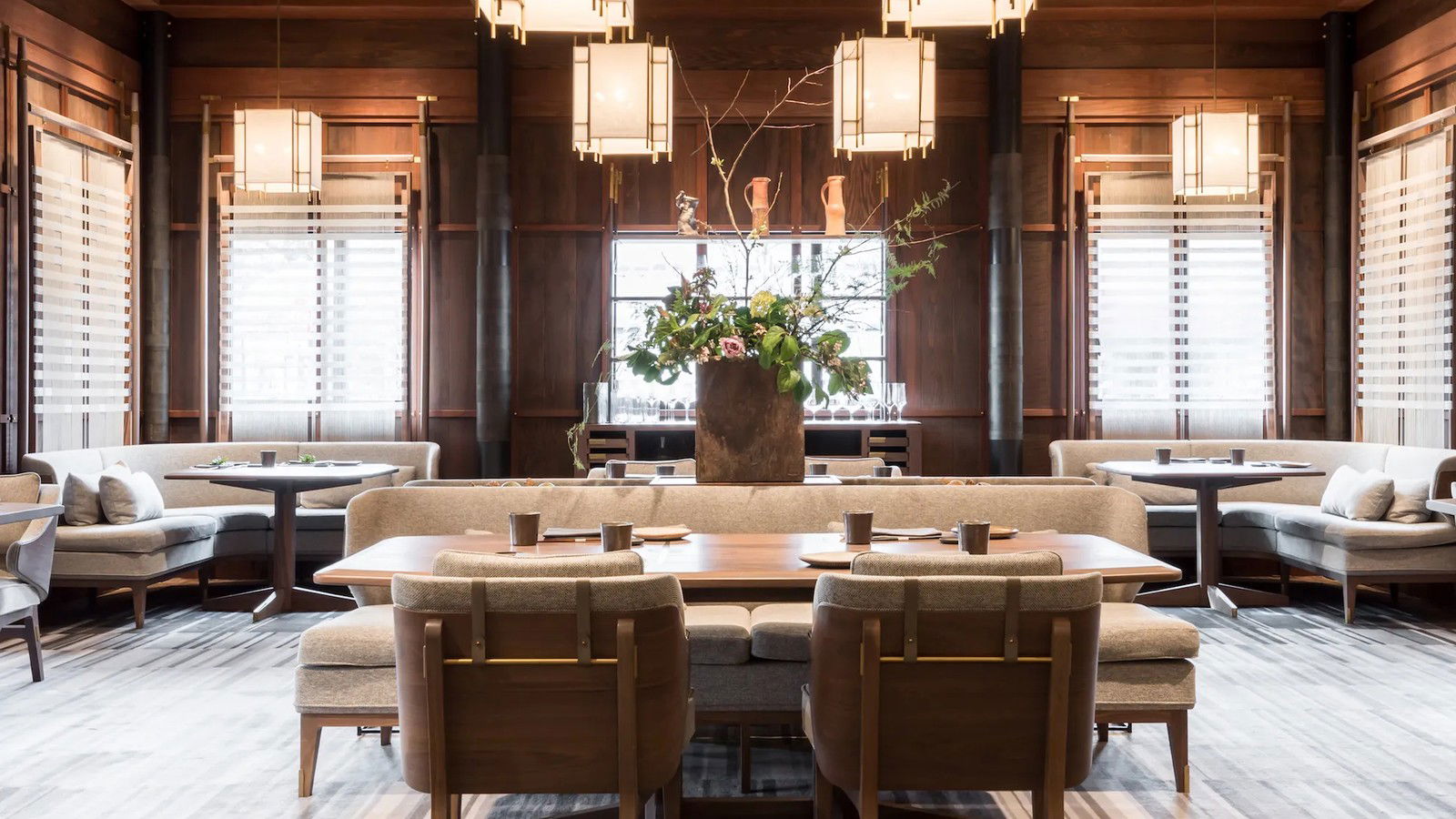 A dining room with dark wood paneling, upholstered banquettes, and a large central table with a floral arrangement under custom pendant lights.