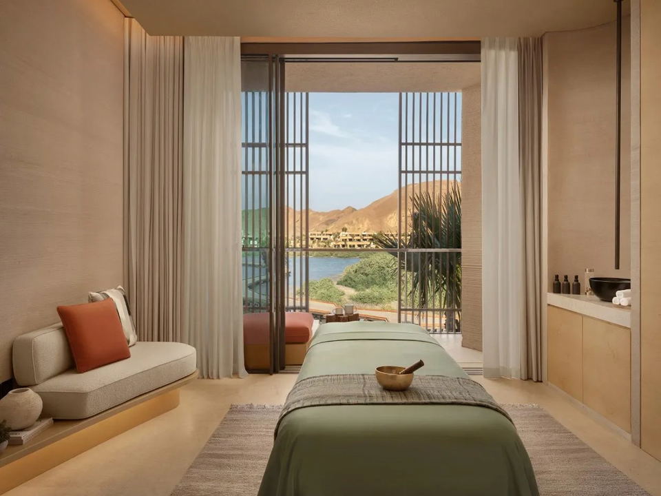 A spa treatment room at Six Senses Amaala with a massage table, a daybed, and an open terrace overlooking a lagoon and mountains.