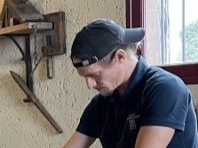 A man wearing a dark cap and black shirt looks closely at wooden barrel-making or cooperage tools mounted on a light-colored wall.