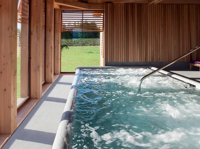 An indoor hydrotherapy pool with bubbling water jets inside a warm cedar-clad spa pavilion, with large windows overlooking a green lawn.