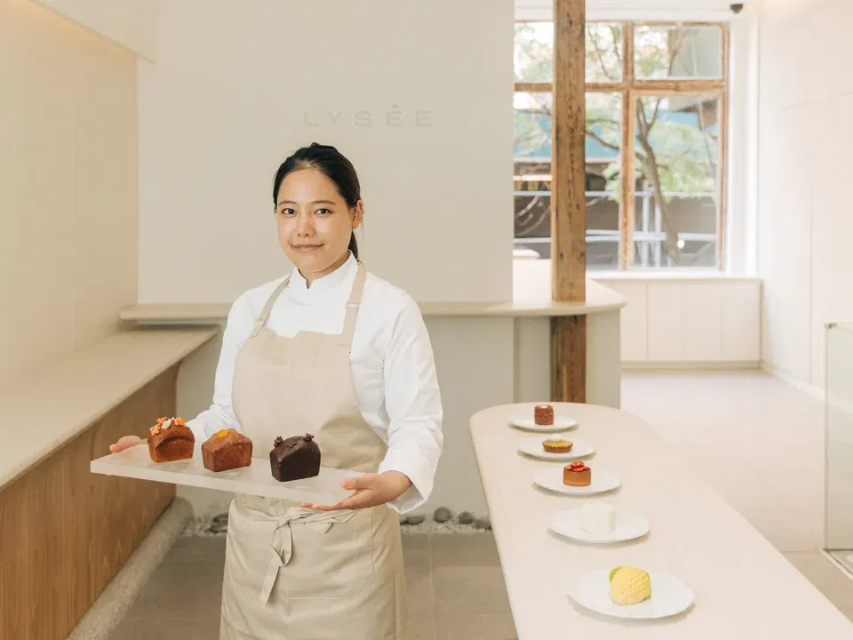 Pastry chef Eunji Lee in a white chef coat and beige apron holds a marble tray with three small cakes inside Lysée, her minimalist New York bakery, with additional plated pastries on the counter behind her.