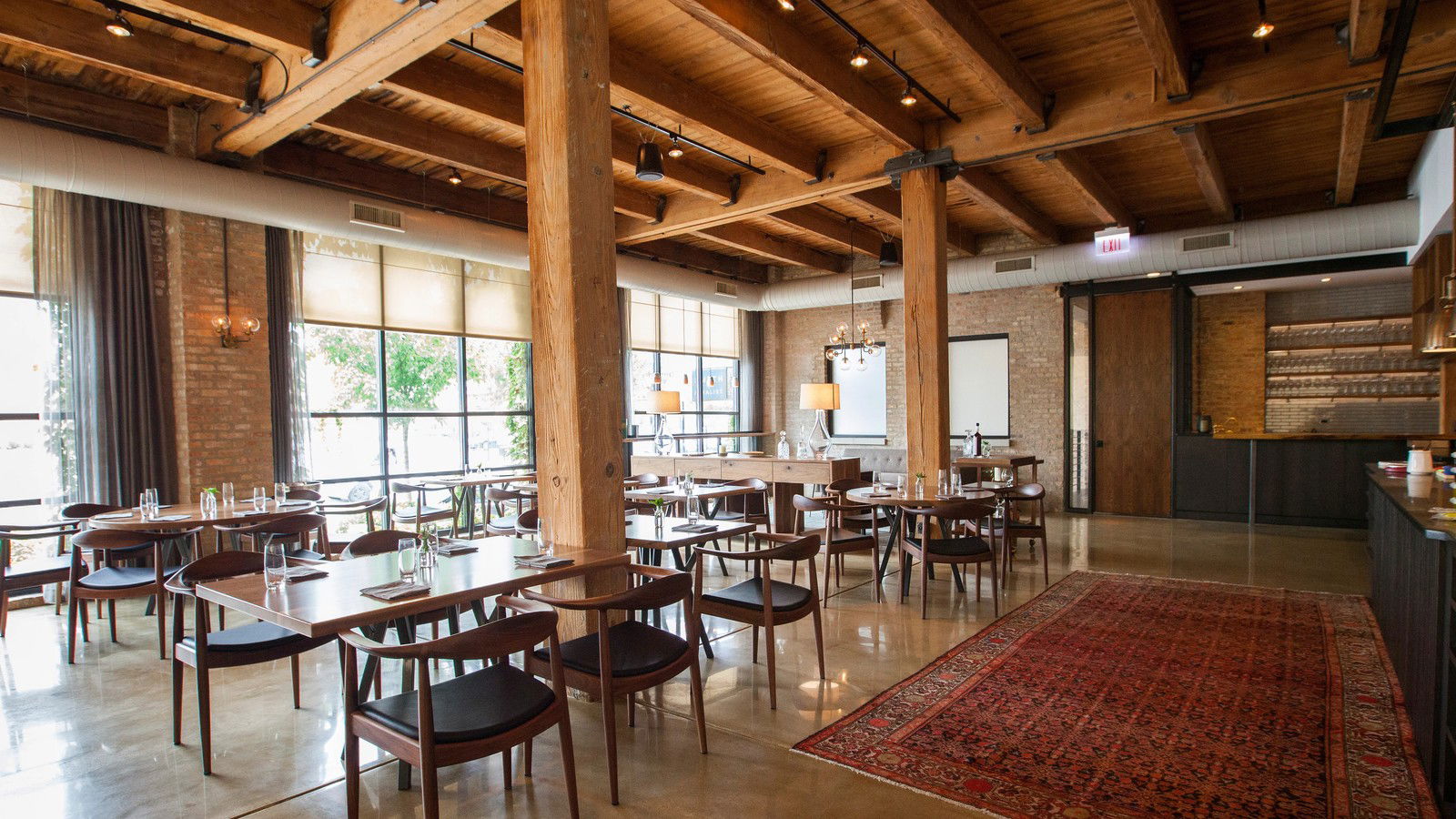 A dining room with exposed wooden beams, brick walls, and tables set with chairs, featuring an oriental rug.