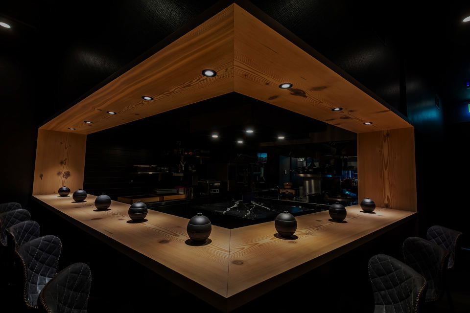 A modern restaurant interior with a U-shaped wooden counter and black quilted chairs, featuring small black decorative urns on the counter.