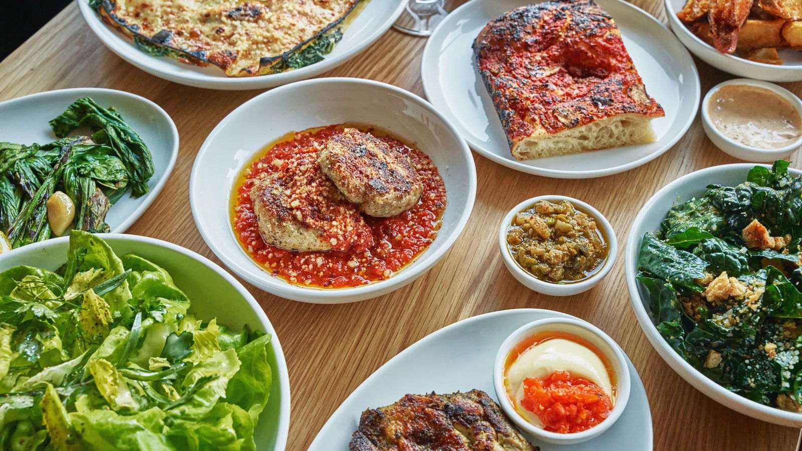 An overhead shot of multiple dishes spread across a wooden table, including flatbreads, meatballs in tomato sauce, and salads.