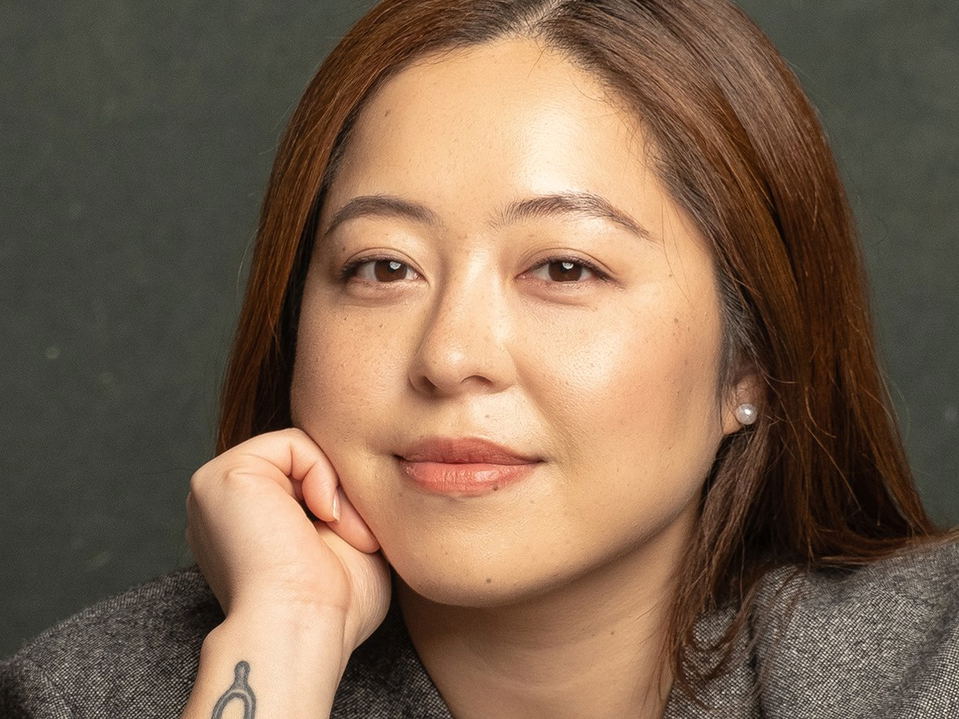Ahris Kim, with reddish-brown hair and a grey blazer, rests her chin on her hand, revealing a wishbone tattoo on her wrist.