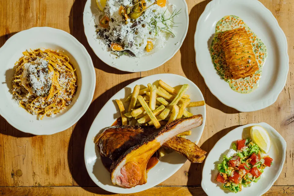 An overhead shot of five dishes on a wooden table, including pasta, salad with cheese, a potato dish with roe, ribs with fries, and a tuna salad.