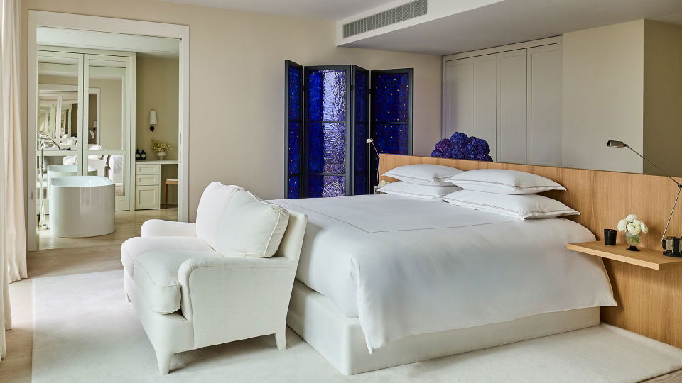 A luxury hotel suite bedroom with white bedding, cream armchairs, a blue decorative glass screen divider, and an en-suite bathroom visible through doo