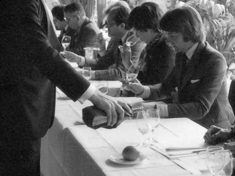 A waiter in a bow tie pours wine for judges seated at a long table during a formal tasting event, with multiple wine glasses and small plates visible.