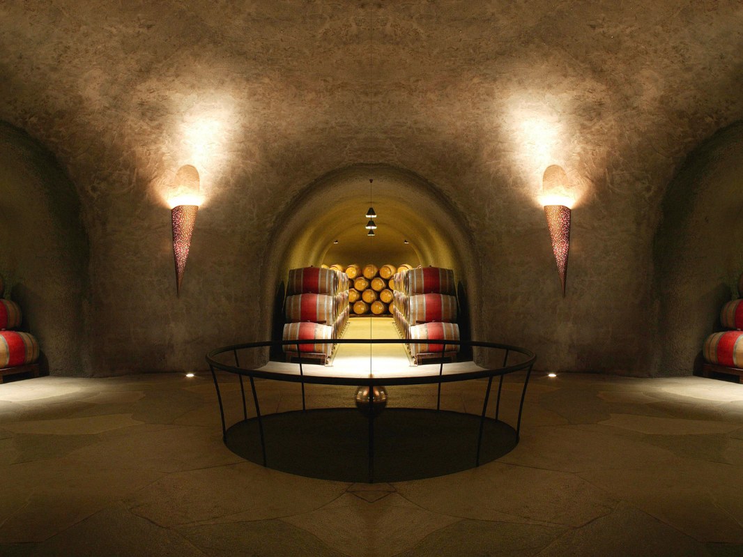 An underground wine cave with arched stone ceilings, wall sconces, and rows of oak barrels stacked in alcoves.