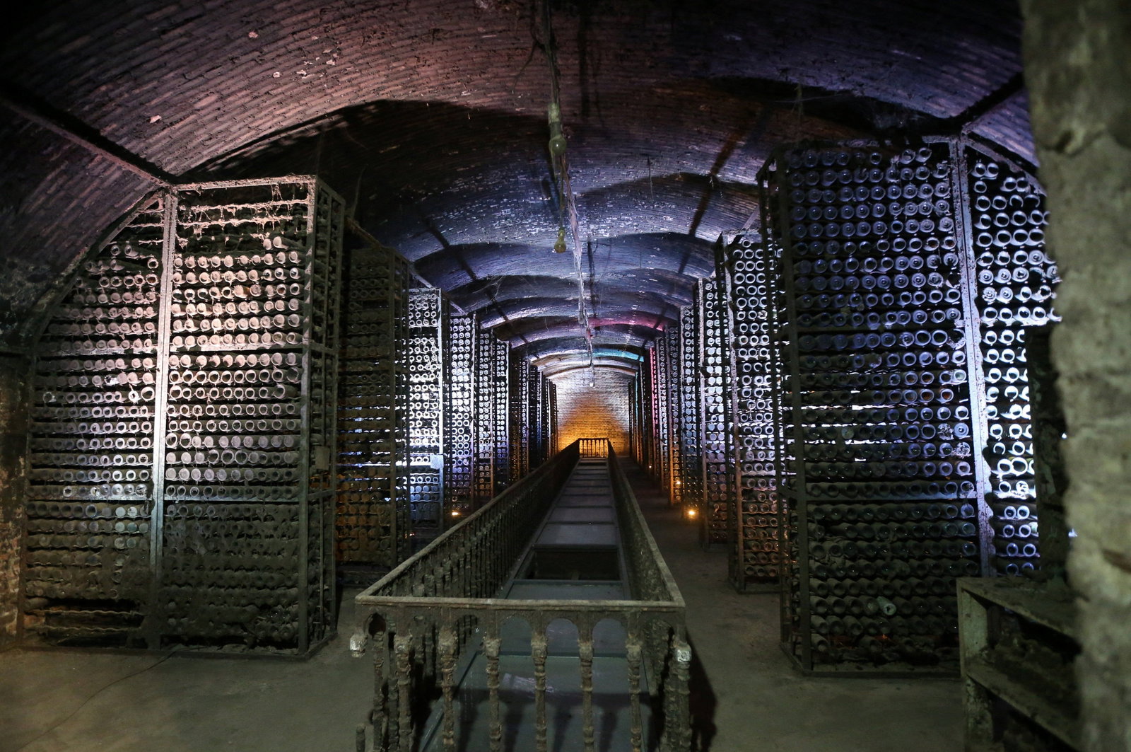 A long, dark, arched cellar with thousands of wine bottles stacked in metal racks along both sides, leading to a distant light.