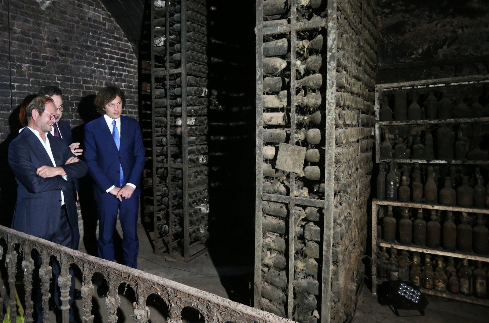 Three men in suits stand in a dark, brick-walled cellar filled with dusty, old wine bottles on metal shelves. The bottles are covered in a thick