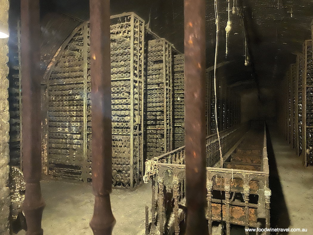 A dark, dusty wine cellar with rows of stacked wine bottles in metal racks, and a long, narrow staircase leading into the distance.
