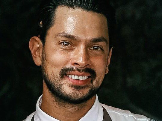 A smiling man with slicked-back dark hair, mustache, and beard, wearing a white chef shirt and dark grey apron, photographed in a doorway.