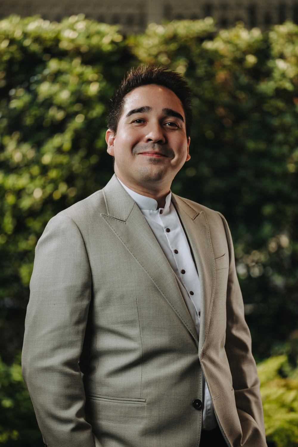 Professional headshot of a man wearing a tan blazer over a white dress shirt, photographed outdoors with green hedges behind him.