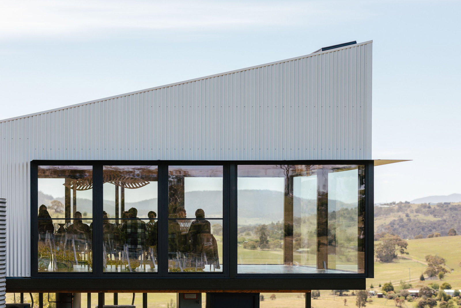 A modern building with a corrugated metal facade and large glass windows, revealing people seated inside with a scenic view.