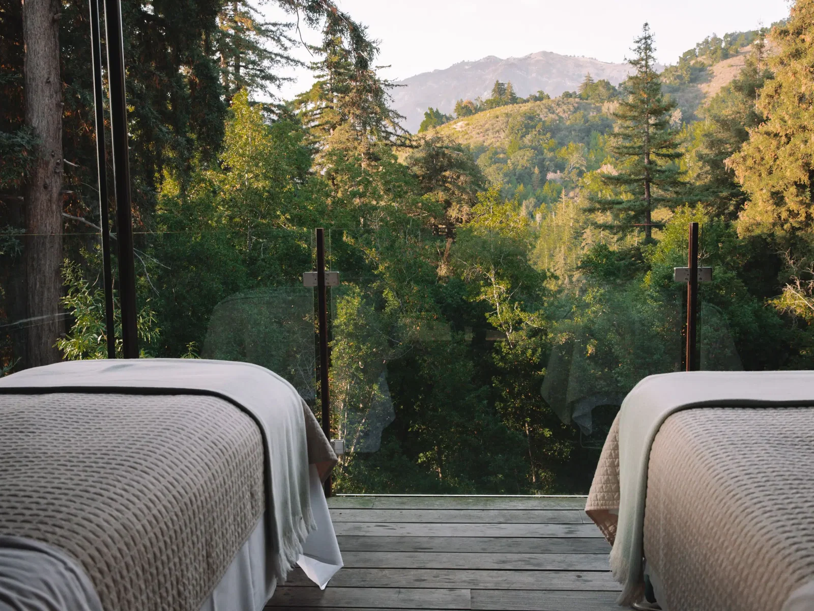 Two massage beds on a wooden deck overlook a lush green forest and distant mountains under a bright sky.