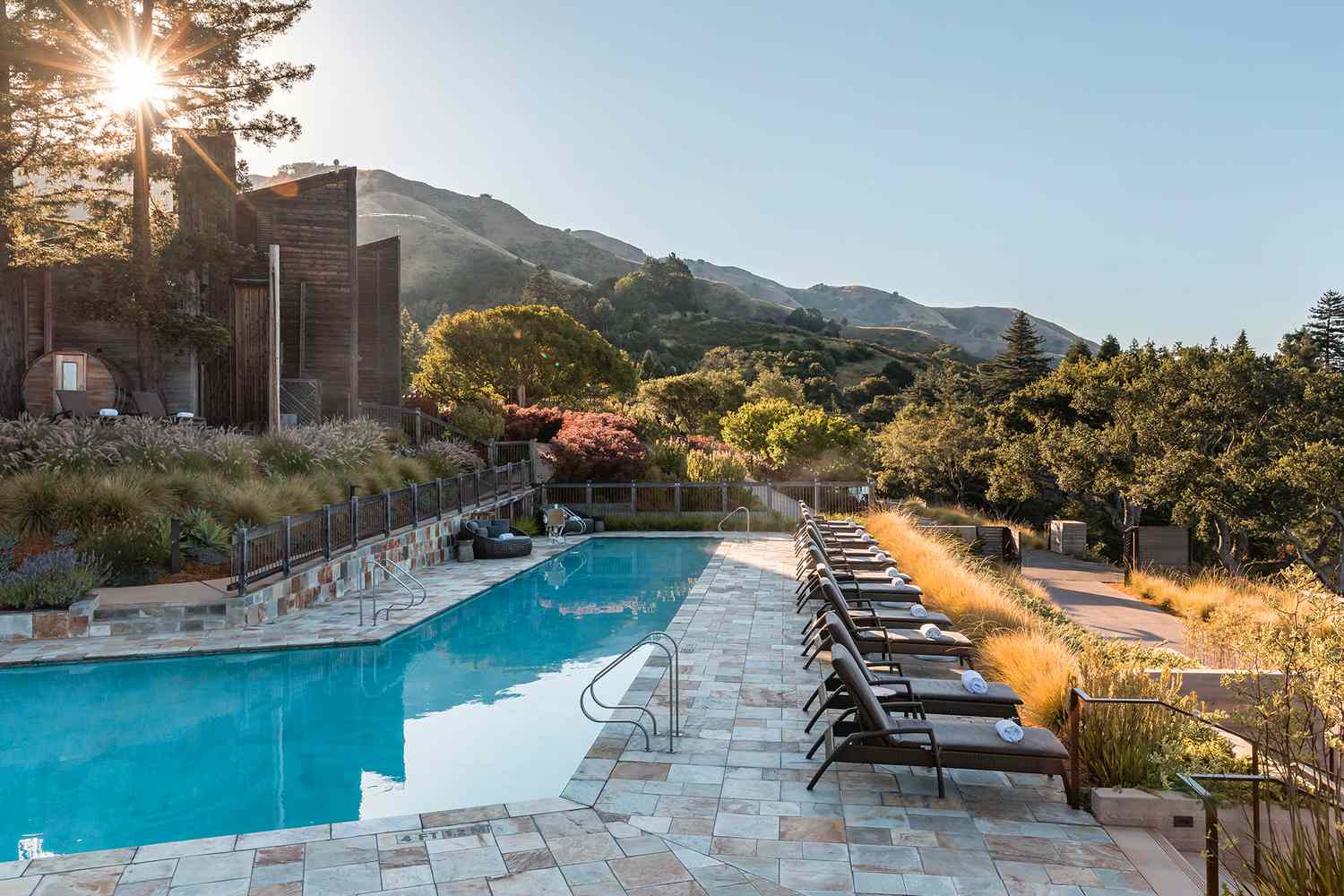 An outdoor swimming pool with lounge chairs at Alila Ventana Big Sur Hotel, surrounded by lush greenery and mountains under a bright sky.