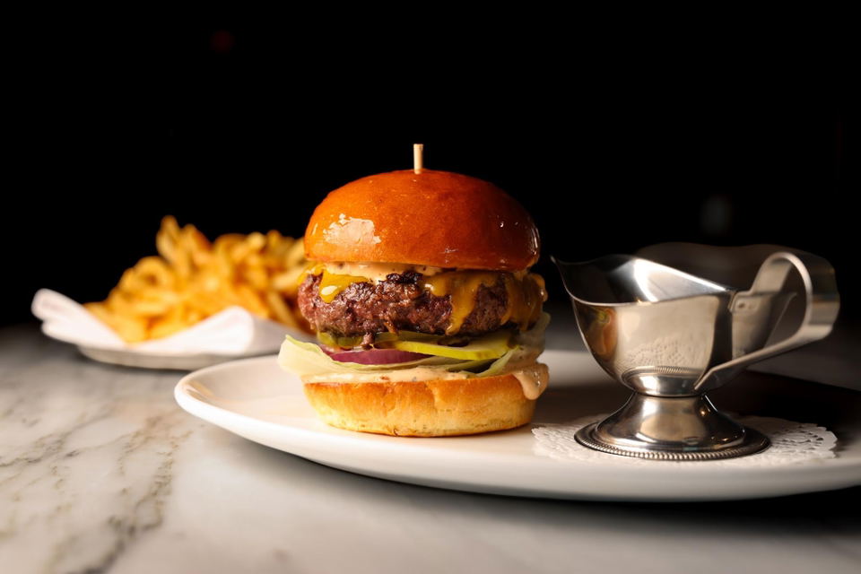A professionally photographed burger with melted cheese, pickles, and onions on a brioche bun, served with fries and a sauce boat on a white plate; th