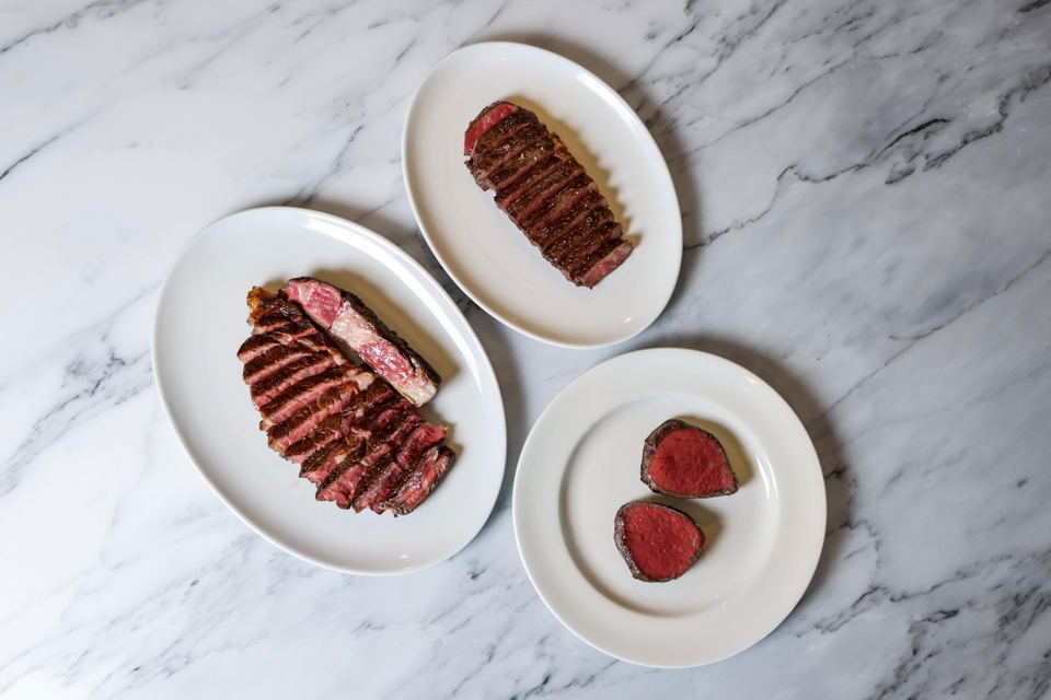 Three white plates on marble surface displaying sliced steaks cooked to various temperatures, presented in an editorial food photography style; source