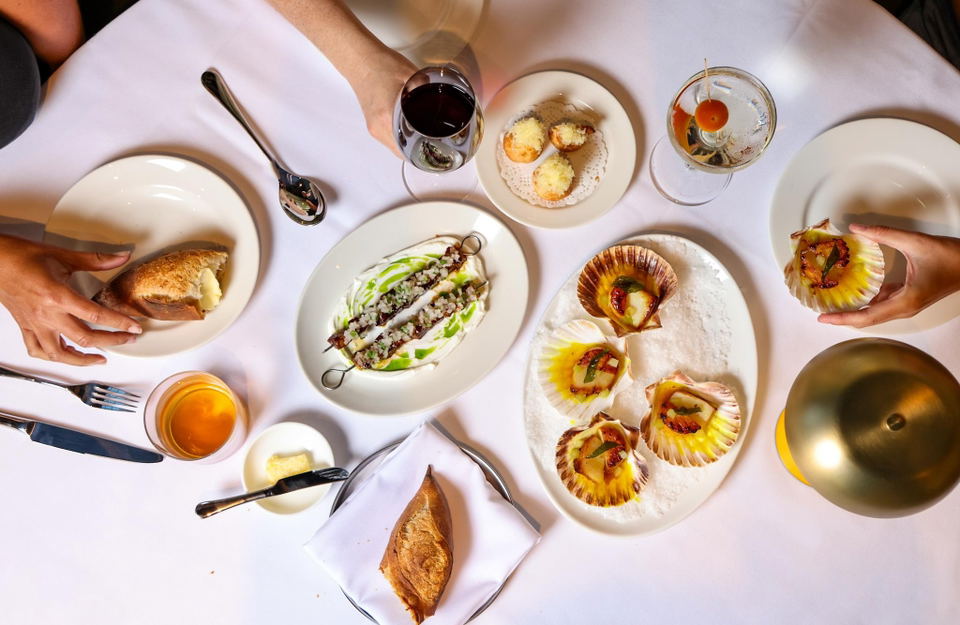 An overhead shot of a restaurant table spread featuring multiple dishes including scallops in shells, gougères, bread, wine glasses, and cocktails; th