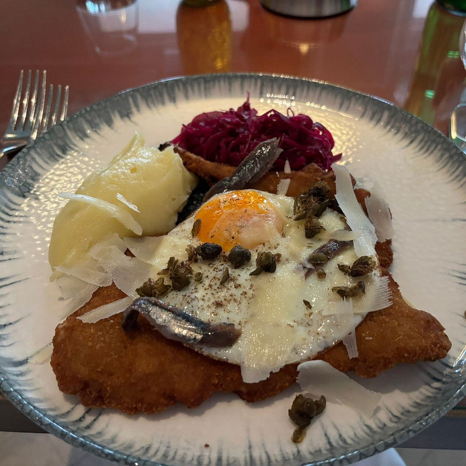 Golden breaded pork schnitzel on a patterned ceramic plate, topped with a fried egg, anchovies, capers, and shaved cheese, with mashed potato and red cabbage.