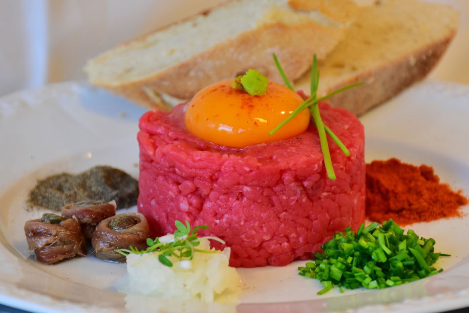 A close-up of a steak tartare dish, featuring a mound of raw minced beef topped with a bright orange egg yolk and a small dollop of green relish.