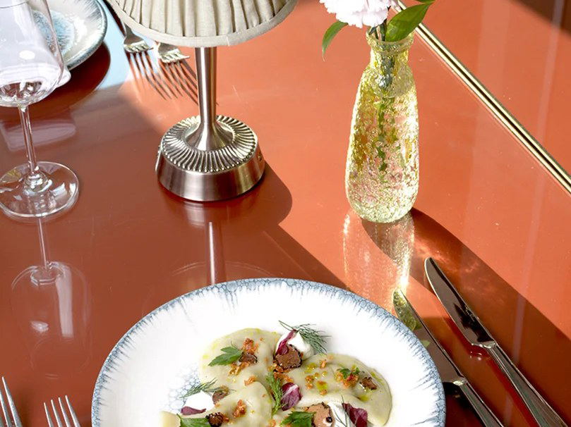 An elegantly plated pasta dish with herbs, truffle shavings, and garnishes on a decorative plate, set on a glossy terracotta-red table with silver