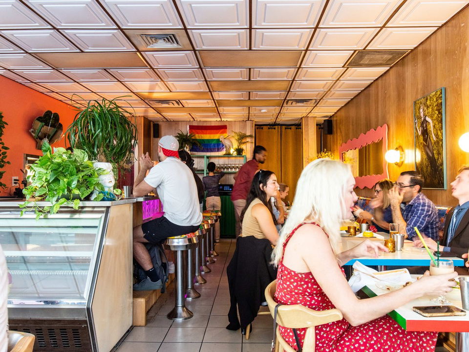 The interior of Tâm Tâm restaurant in Miami, with patrons seated at a counter and tables, and a rainbow Pride flag hanging in the background.