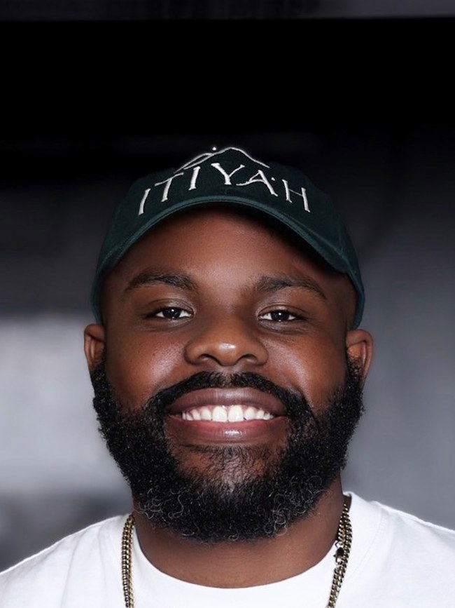 A smiling bearded man in an Itiyah cap, likely chef Sebastien Salomon, in a professional kitchen setting.
