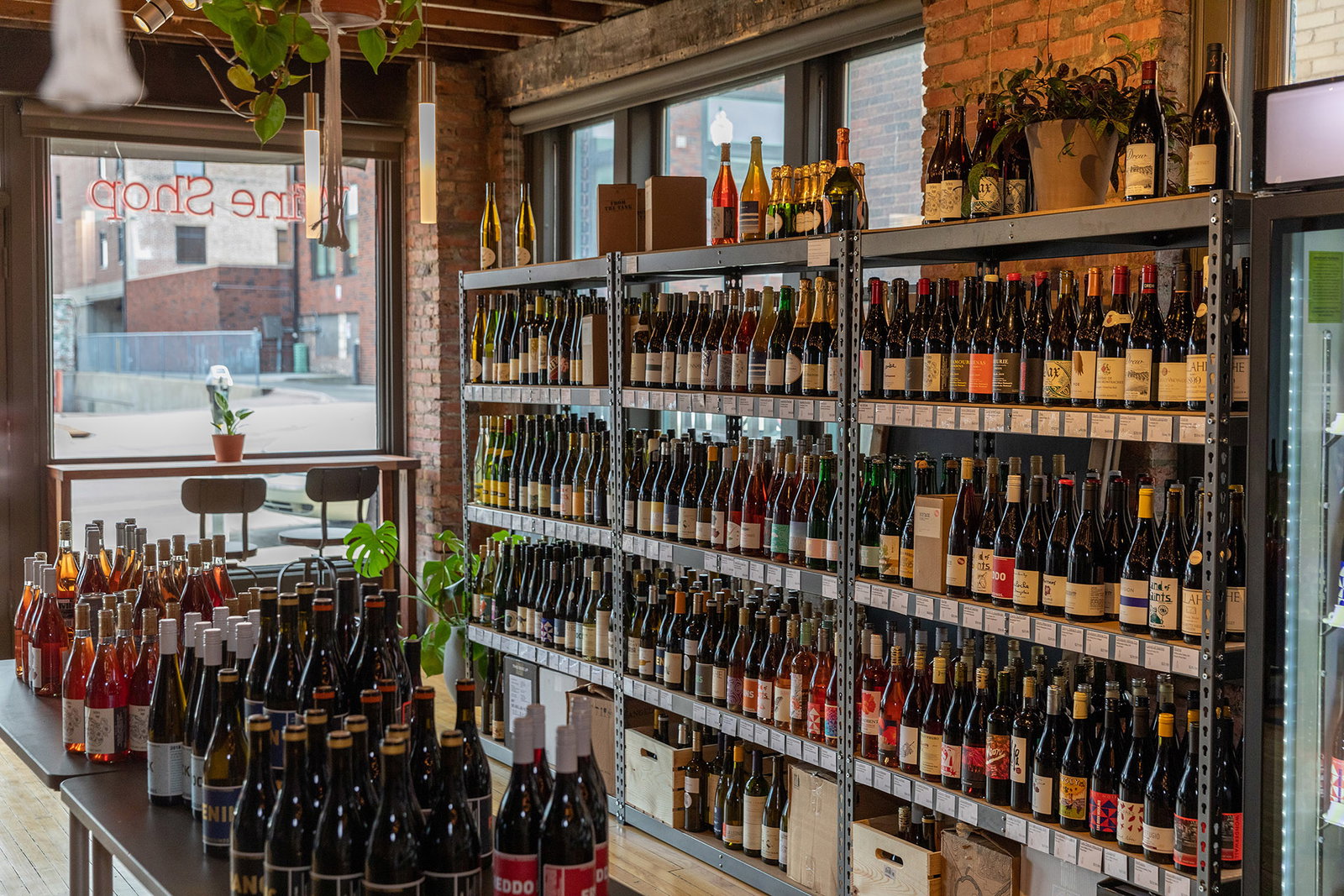 Interior of a wine retail shop showing extensive metal shelving filled with wine bottles, a display table with bottles in the foreground, exposed bric