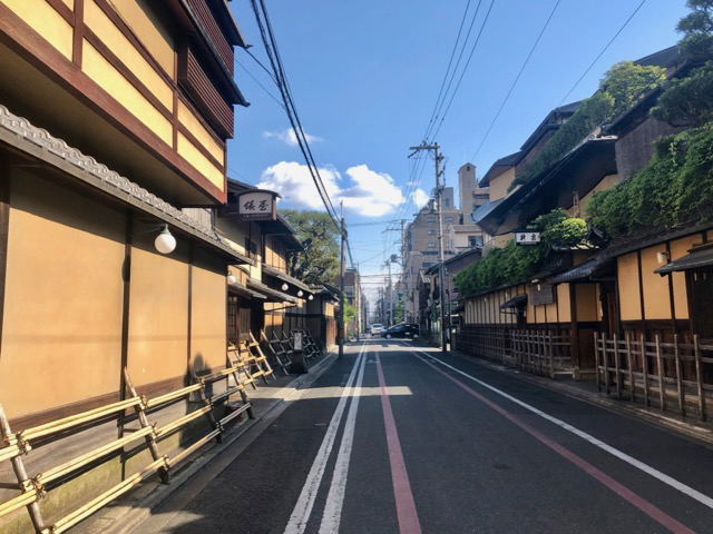 A narrow street in Japan with traditional wooden buildings on both sides, power lines overhead, mountains in the background, and clear blue sky.
