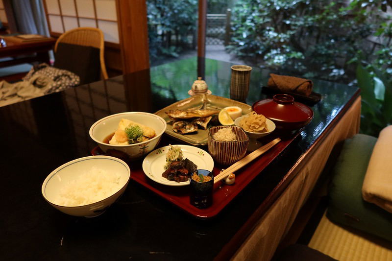 A formal Japanese meal setup on a black table featuring a red lacquered tray with various small ceramic bowls containing different dishes, a rice bowl, gold-trimmed cups, and traditional serving vessels, with a garden view visible through windows in the background.
