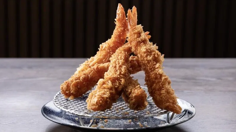Panko-fried shrimp, known as ebi fry, are stacked on a wire rack over a dark ceramic plate, showcasing their crispy, golden-brown coating.