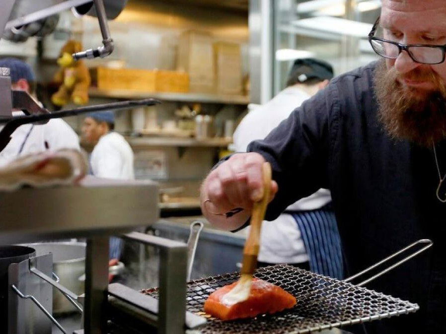 A bearded chef with glasses brushes a white glaze onto a salmon fillet on a wire rack over a grill in a professional kitchen.