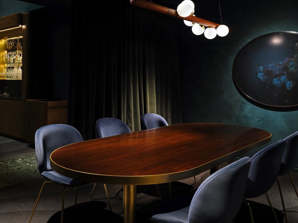 A dark, moody dining room with an oval walnut table, blue velvet chairs, a sculptural pendant light, teal walls, and a bar.