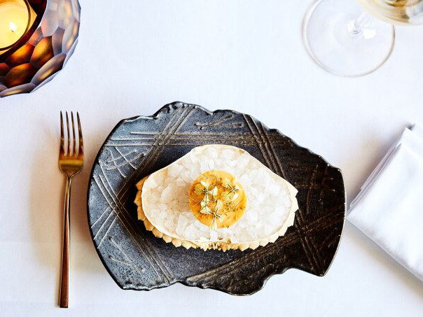 An overhead close-up of an elegantly plated dish on a dark textured ceramic plate, accompanied by a gold fork, wine glass, candle, and folded napkin