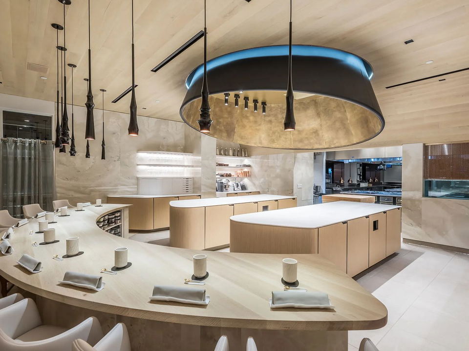A modern fine-dining restaurant interior with a curved light wood chef's counter, dramatic black pendant lights, and an open kitchen.