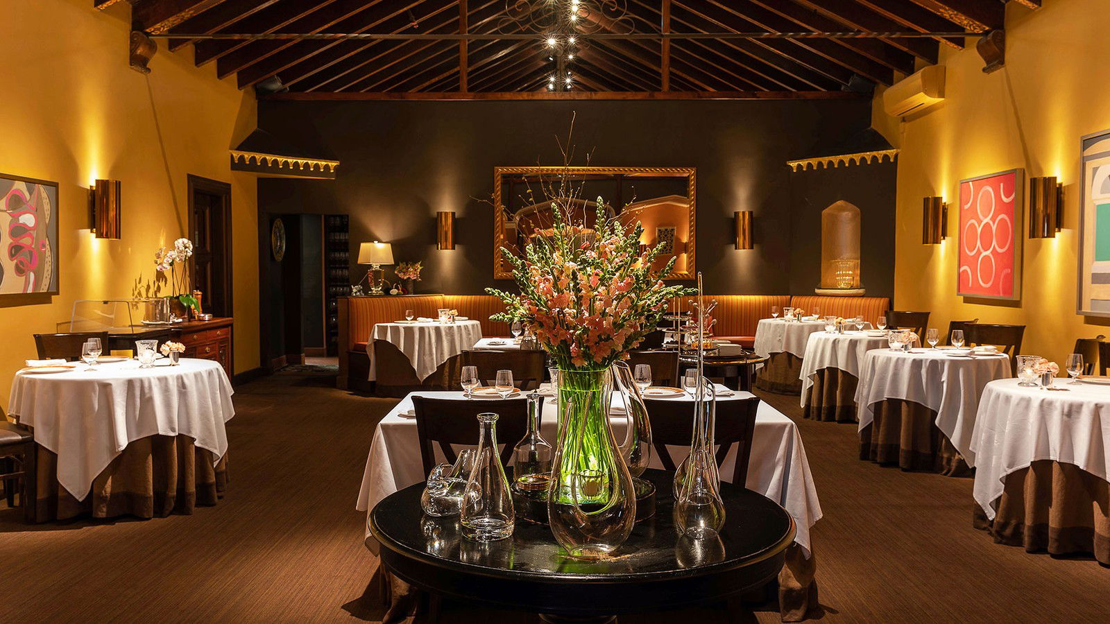 An elegant, warmly lit fine-dining room interior with white-tablecloth tables, exposed dark wood ceiling beams, and artwork.
