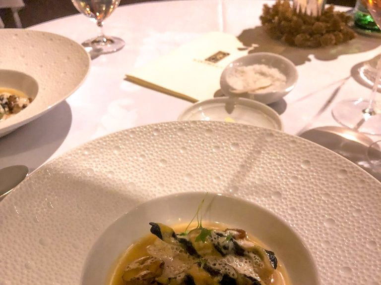 A close-up of an elegantly plated pasta or ravioli dish with mushrooms and foam sauce on a textured white plate at a formal restaurant table.