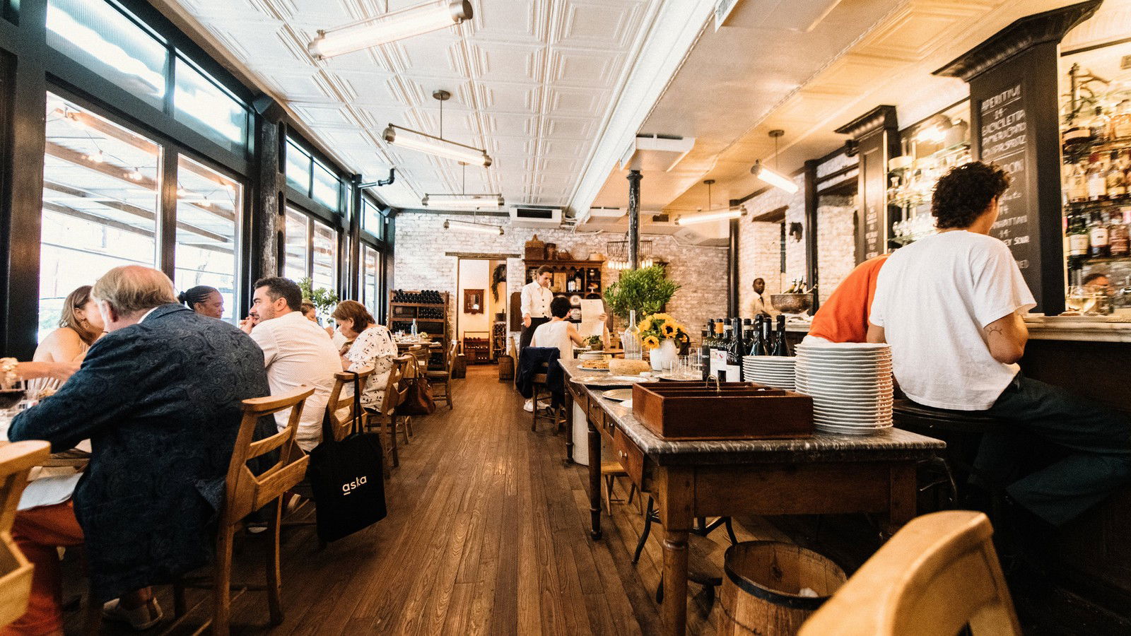 A bustling Italian trattoria dining room with a tin ceiling, large windows, wooden tables and chairs, and a bar area.